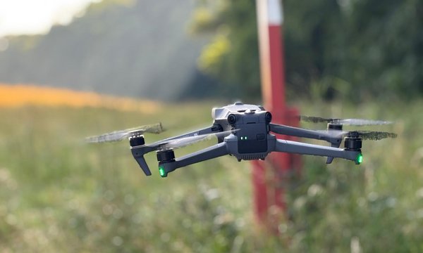 Eine graue Drohne schwebt in der Luft auf einer Wiese mit unscharfem Hintergrund aus Bäumen und einem rot-weißen Pfosten.