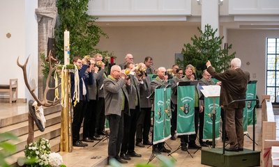 Eine Bläsergruppe spielt Jagdhörner unter der Leitung eines Dirigenten in einem großen, geschmückten Innenraum mit Pflanzen und Dekorationen. Text auf grünen Bannern: "Jägertarnung Bayern e.V. Jagdhornbläserkorps".