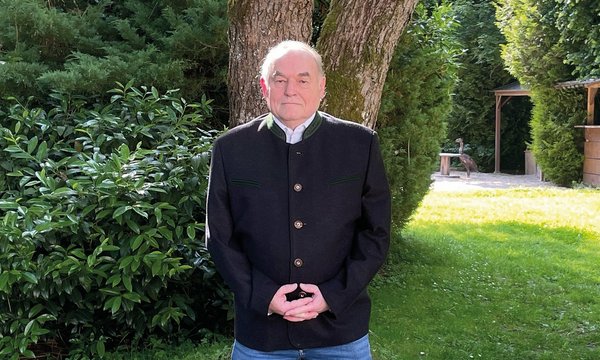 Ein älterer Mann steht ruhig in einem gepflegten Garten, trägt eine dunkle Jacke und blickt geradeaus. Im Hintergrund sind Sträucher und ein großer Baum sichtbar.